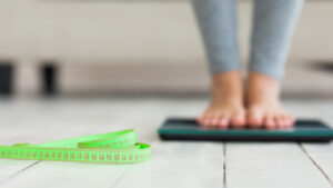 Picture of a woman standing on a bathroom scale with a tape measure on the ground next to her.