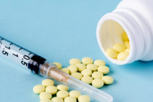 Picture of a syringe among yellow pills spilling out from a white bottle.