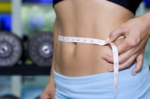 Picture of a woman holding a tape measure around her waist with hand weights in the background.