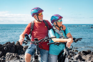 Senior couple of cyclists wearing helmets resting on a beach 